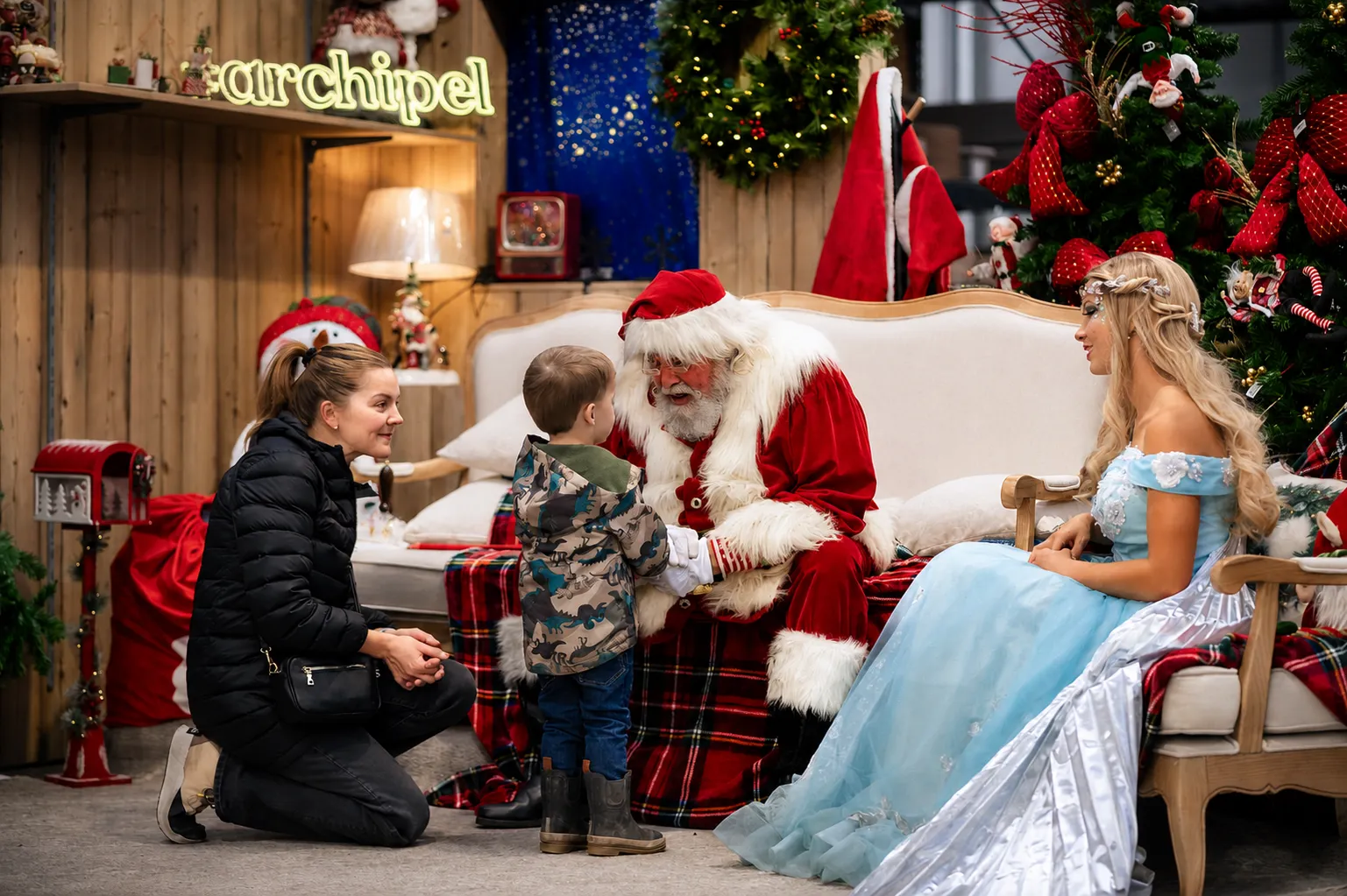 Le Père Noël, assis sur une causeuse, accueille un petit garçon à côté d'une fée en robe bleue, sous l'enseigne de l'Archipel.