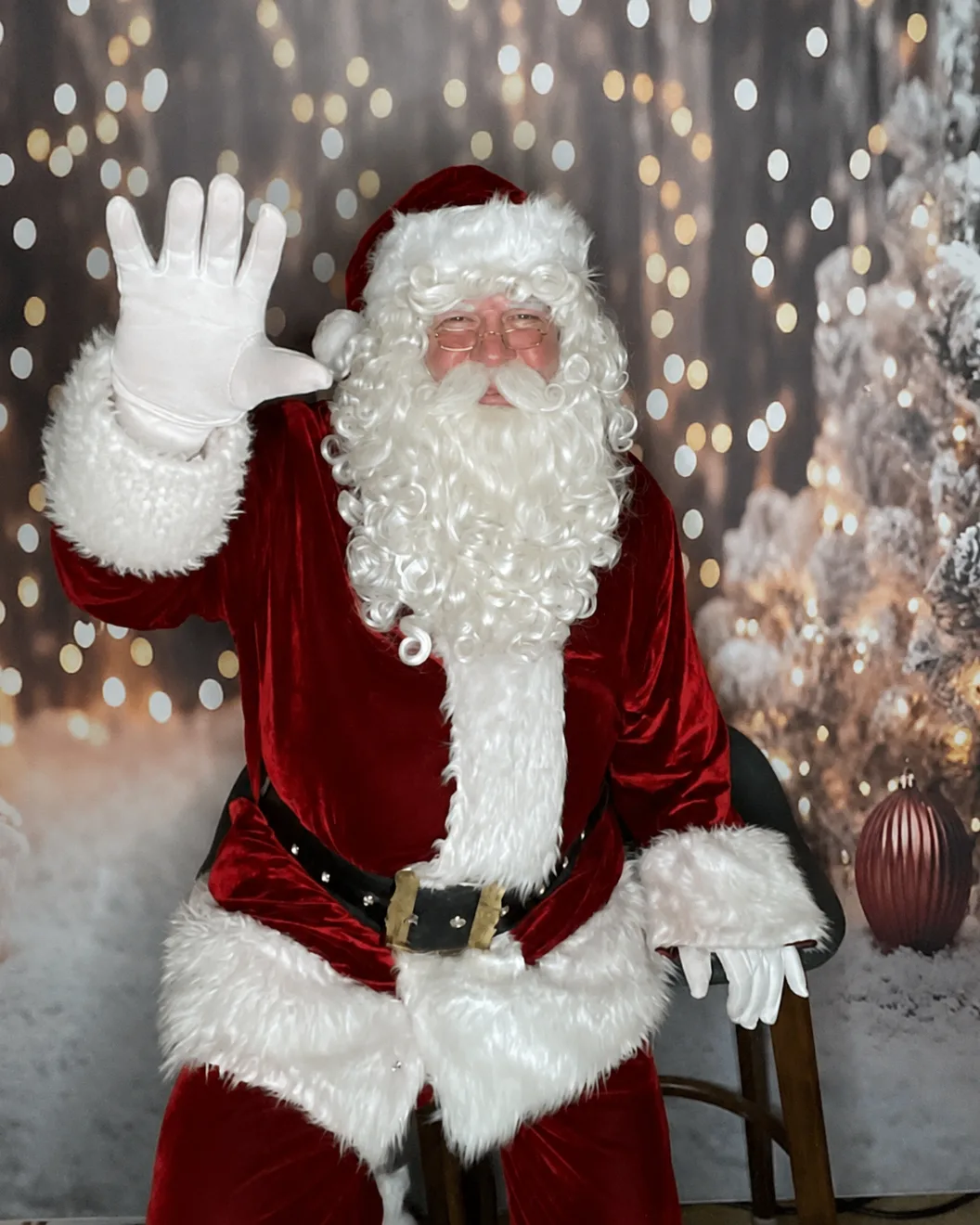 Le Père Noël Michel, la main levée pour saluer, devant un décor de forêt enneigée illuminée.